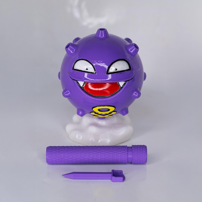 May include: A purple, spherical figurine with a smiling face, white teeth, and a red tongue. It sits atop a white cloud base, with a matching purple cylindrical piece and a pointed tool below. The figurine has a yellow and black symbol.