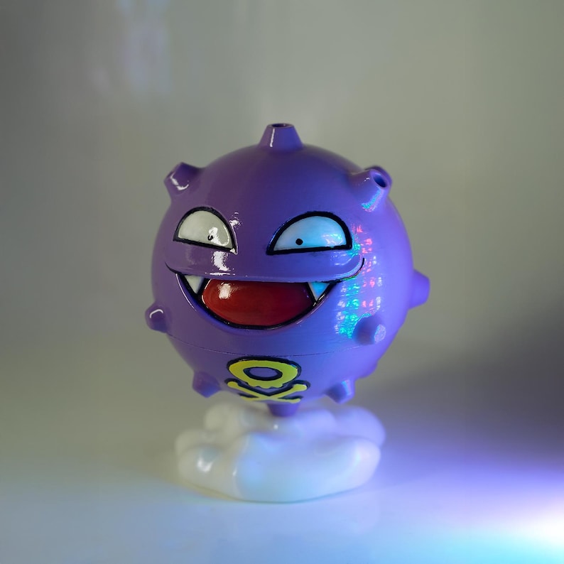 May include: A purple, spherical figurine with a smiling face, sharp teeth, and a red mouth. The figurine has a yellow and black skull and crossbones symbol on its base. It is perched on a white, cloud-shaped base.