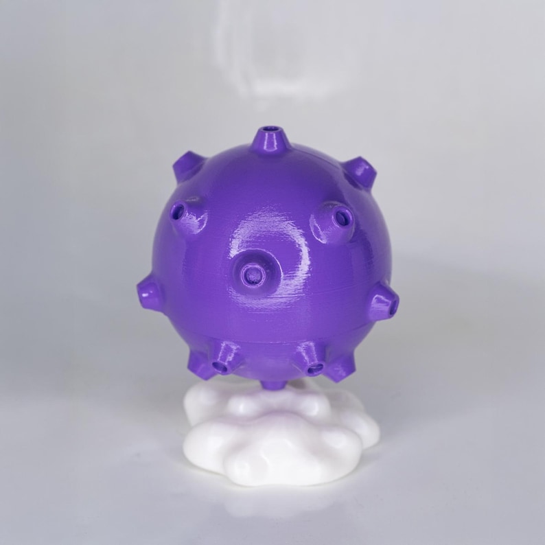 May include: A purple, spherical object with protruding spikes and circular indentations, resting on a white cloud-shaped base. The object has a glossy finish and appears to be a decorative item or a unique piece of art.