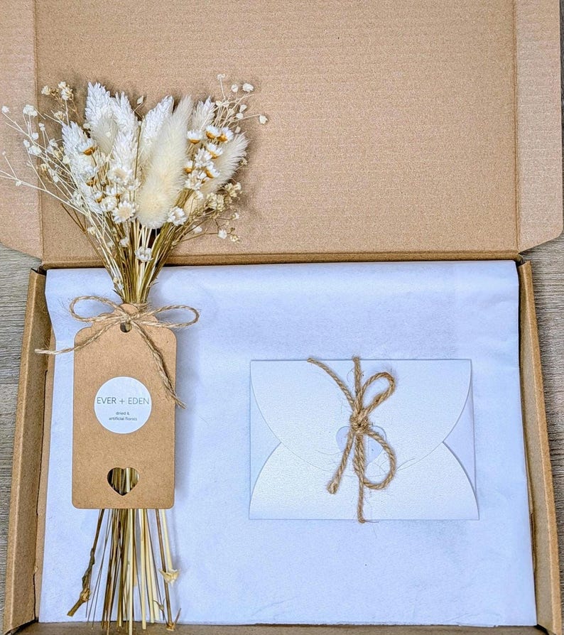 May include: A brown gift box containing a bouquet of dried flowers, a tag with the text "EVER + EDEN", and a white card tied with twine. The contents are arranged on white tissue paper.