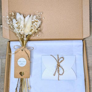 May include: A cardboard box containing a bouquet of dried flowers, a gift tag, and a white envelope tied with twine. The bouquet features white and beige flowers. The gift tag reads "EVER + EDEN".