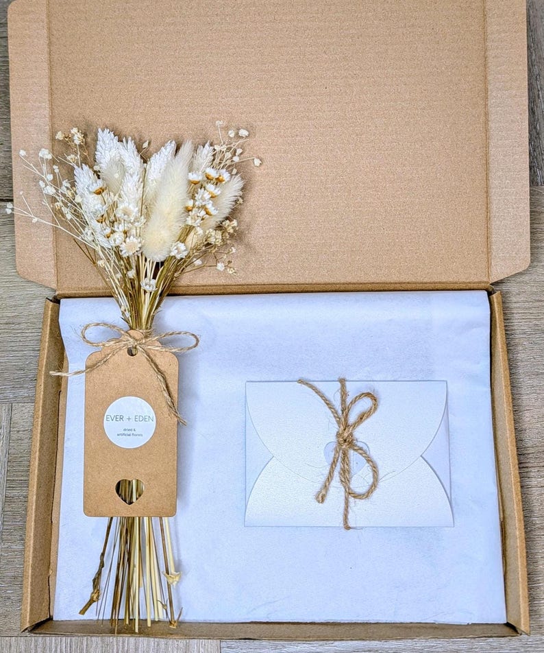 May include: A cardboard box containing a bouquet of dried flowers, a gift tag, and a white envelope tied with twine. The bouquet features white and beige flowers. The gift tag reads "EVER + EDEN".