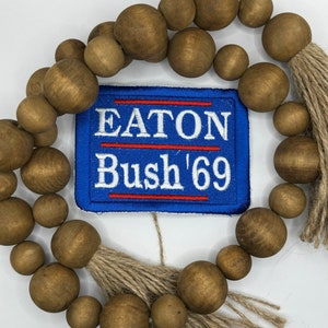 May include: A wooden bead garland with brown wooden beads and jute tassels. The garland has a blue and red patch with the text "EATON Bush '69" in white.