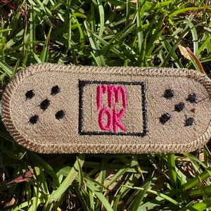 May include: A beige fabric patch shaped like a bandage with black stitching around the edges. The patch has black embroidered dots and a black square with the words "I'm OK" embroidered in pink.