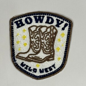 May include: Embroidered patch with a shield shape, featuring a pair of cowboy boots in brown and white. The patch has the words "HOWDY!" and "WILD WEST" in navy blue, with yellow star accents. The border is navy blue.