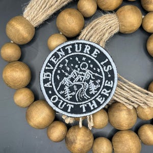 May include: Black and white embroidered patch with a mountain scene and the text "Adventure is out there". The patch is surrounded by wooden beads.