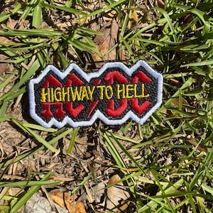 May include: Embroidered patch with the text "HIGHWAY TO HELL" in red and yellow lettering. The patch has a black background with a white border. The patch is rectangular and is set against a background of green grass.