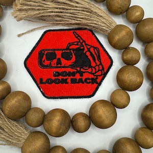 May include: A red hexagonal patch with a black skeleton hand pointing to a black and white image of a phone with the text "Don't Look Back". The patch is surrounded by a brown wooden bead garland.