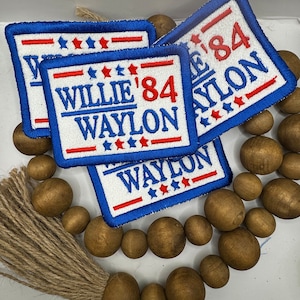 May include: Three blue and red embroidered patches with white text that reads "Willie '84 Waylon". The patches are on a brown wooden beaded garland with a brown tassel.