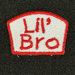 May include: White embroidered patch with a red outline. The patch says "Lil' Bro" in red lettering.
