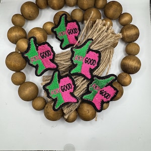 May include: A wooden bead garland with five pink and green embroidered patches that say "For Good" in black lettering.