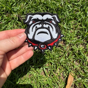 Peut inclure: Patch brodé blanc représentant un bouledogue avec un collier rouge et noir. Le bouledogue a une expression féroce avec des yeux noirs et un nez noir. Le collier a un 'G' noir et blanc au centre.
