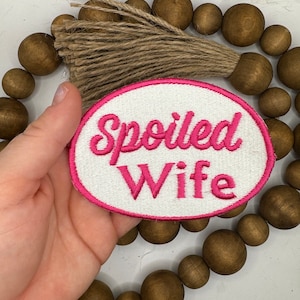 May include: A white, round patch with a pink border. The text "Spoiled Wife" is embroidered in pink on the patch.