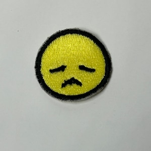May include: Yellow embroidered iron-on patch with a sad face. The face has closed eyes and a small mustache.