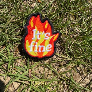 May include: A red, orange, and yellow embroidered patch shaped like a flame with the text "It's fine" in white.