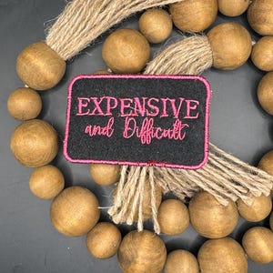 May include: A black fabric patch with pink embroidery that reads "Expensive and Difficult" on a background of brown wooden beads with jute tassels.