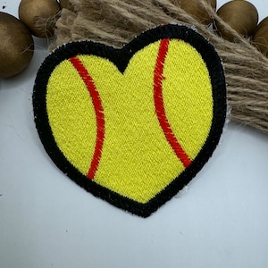 May include: A yellow heart-shaped patch with a red softball design. The patch has a black outline.