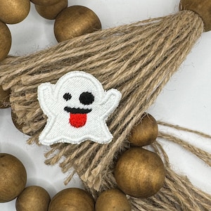 May include: A white ghost with a black eye and a red tongue is embroidered on a brown burlap tassel. The tassel is attached to a string of brown wooden beads.