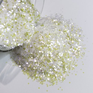 May include: A clear glass jar tipped over, spilling a mix of white and iridescent glitter with specks of light green glitter onto a white surface. The glitter is in various shapes and sizes.