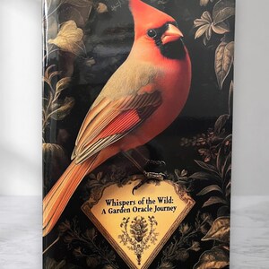 Op de afbeelding: Een hardcover boek getiteld Whispers of the Wild: A Garden Oracle Journey, met een felrode kardinaalvogel op een goudomrand label tegen een donkere achtergrond met goudkleurige botanische illustraties van bladeren en bloemen.