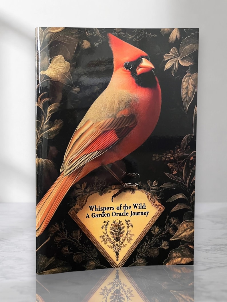 Op de afbeelding: Een hardcover boek getiteld Whispers of the Wild: A Garden Oracle Journey, met een felrode kardinaalvogel op een goudomrand label tegen een donkere achtergrond met goudkleurige botanische illustraties van bladeren en bloemen.