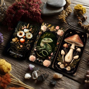 Op de afbeelding: Drie rechthoekige tarotkaarten met een zwarte achtergrond. De linkerkaart toont witte bloemen en een oranje vrucht met de tekst Embrace the Wild. De middelste kaart toont een groene hagedis omringd door roze rozen met de tekst Wisdom. De rechterkaart toont paddenstoelen met de tekst Growth.