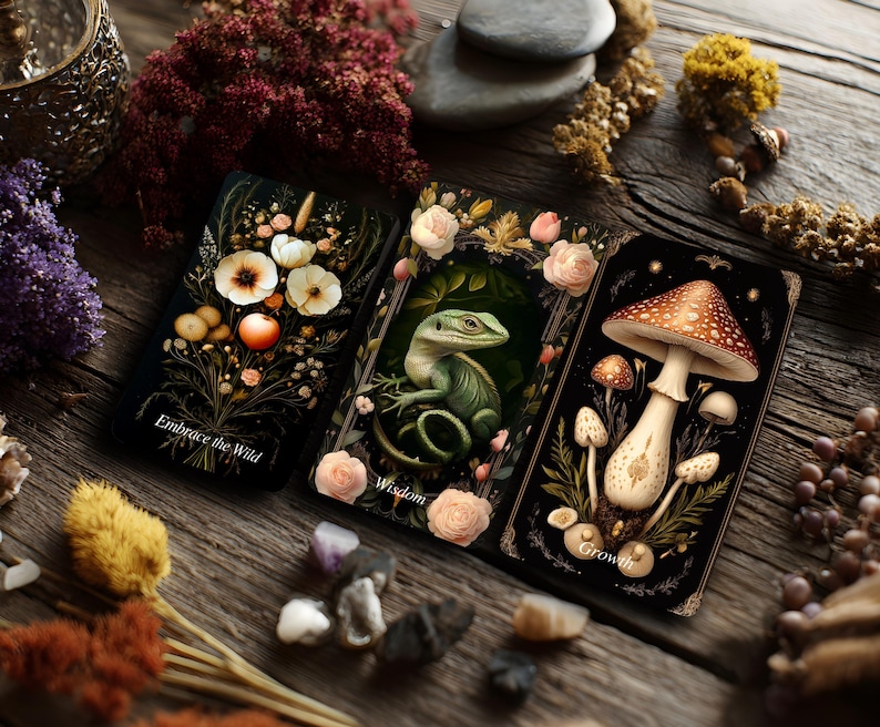 Op de afbeelding: Drie rechthoekige tarotkaarten met een zwarte achtergrond. De linkerkaart toont witte bloemen en een oranje vrucht met de tekst Embrace the Wild. De middelste kaart toont een groene hagedis omringd door roze rozen met de tekst Wisdom. De rechterkaart toont paddenstoelen met de tekst Growth.