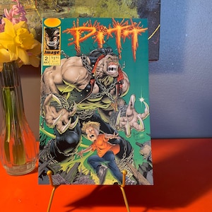 May include: A comic book cover featuring a green, red, and black illustration of a muscular, clawed creature with a chain around its neck. The creature is standing over a smaller, blonde-haired character. The title "Pitt" is written in large, red letters across the top of the cover. The comic book is labeled "Issue #2, July".