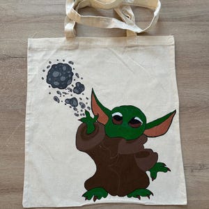 Peut inclure: Un sac fourre-tout en toile beige avec un personnage vert aux grandes oreilles et une cape marron. Le personnage fait un geste vers un amas de roches grises. Le sac a deux poignées beiges.