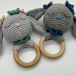 Puede incluir: Dos juguetes de sonajero de conejo de crochet gris con anillos de madera. Un conejo tiene acentos verdes y azules, el otro tiene acentos azules y beige.