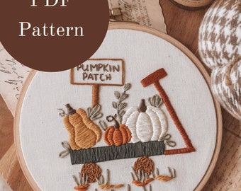 May include: A white embroidery hoop with a brown wooden frame, featuring a hand-stitched design of a pumpkin patch with a wagon full of pumpkins. The design includes a sign that reads "Pumpkin Patch".