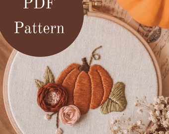 May include: Embroidered hoop art featuring a pumpkin with leaves and flowers. The hoop is on a wooden surface with dried flowers and a small pumpkin.