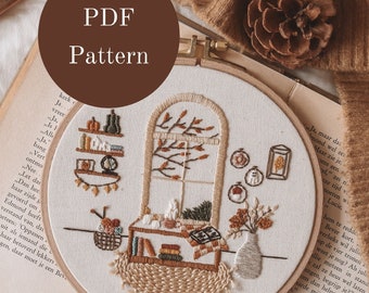 May include: A brown embroidery hoop with a beige fabric inside. The embroidery depicts a window with a bookshelf, a vase with flowers, and a basket with yarn. The hoop is on top of a book with brown pages and text in Dutch. The background includes a brown knitted blanket, a pine cone, and a magnifying glass on a wooden surface.
