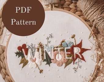 May include: Embroidered hoop art with the word "WINTER" in white letters surrounded by colorful flowers and leaves. The hoop is on a woven mat with dried flowers and twine.