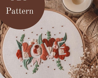 May include: A PDF pattern for an embroidery hoop with the word "LOVE" stitched in red, pink, and white with green leaves and small red dots. The hoop is on a woven brown surface with a candle, twine, and dried flowers.