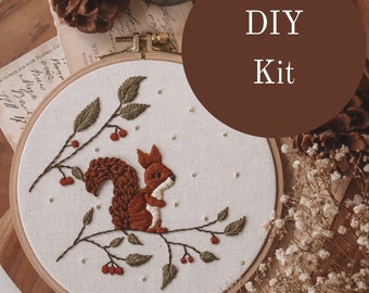 May include: A DIY embroidery kit featuring a brown squirrel on a branch with red berries. The kit includes a hoop, fabric, and embroidery floss. The text "DIY Kit" is written in a circle on a brown background.