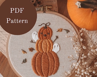 May include: Embroidered hoop art featuring a pumpkin with a face and two ghosts. The pumpkin is stacked on top of two other pumpkins. The hoop is on a wooden surface with dried flowers and a pumpkin.