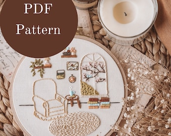 以下が含まれることがあります： 白い刺繍枠に、椅子、テーブル、窓、本のある部屋のカラフルな刺繍模様が施されています。刺繍枠は、籐製のマットの上に置かれ、ドライフラワーとキャンドルが添えられています。