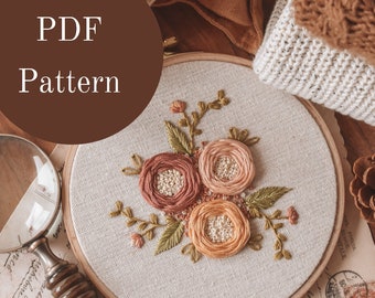 May include: A PDF pattern for embroidery featuring a floral design with three peach and pink flowers and green leaves in a hoop on a wooden surface.