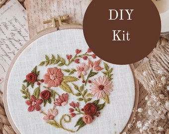 May include: DIY embroidery kit featuring a heart-shaped floral design with pink, red, and green thread on a white fabric hoop. The hoop is surrounded by dried flowers, twine, and a vintage postcard.