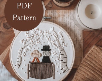 May include: A white embroidery hoop with a design of two people sitting on a bench under a tree with white flowers. The hoop is on a wooden surface with a brown knitted scarf, a candle, and a small vase of white flowers.