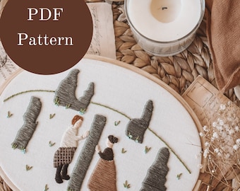 May include: An embroidered hoop art piece featuring two figures and stylized trees. The figures are dressed in brown and tan clothing. The text "PDF Pattern" is in a brown circle. The piece is on a woven mat with a candle and dried flowers.
