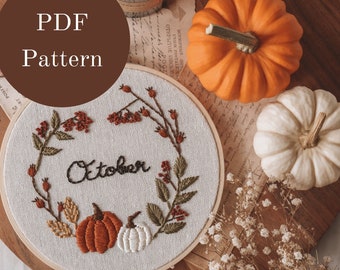 May include: Embroidered hoop art with the word "October" surrounded by fall foliage and pumpkins. The hoop is on a wooden surface with a small bouquet of dried flowers and two pumpkins.