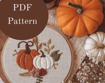 May include: A PDF pattern for an embroidery hoop with a design of three pumpkins, two orange and one white, with leaves and flowers. The hoop is on a wooden surface with a white pumpkin and a bunch of dried flowers.