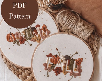May include: Two embroidery hoops with white fabric and brown wooden frames. The fabric is embroidered with the words "Autumn" and "Fall" in colorful thread. The words are surrounded by embroidered leaves, pumpkins, and flowers.