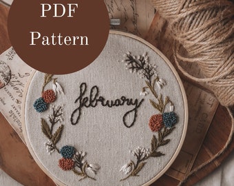 May include: A PDF pattern for an embroidery hoop with the word "February" stitched in black thread and surrounded by a wreath of green leaves and colorful flowers.