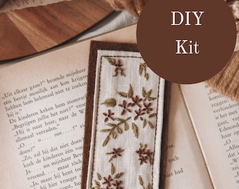 May include: DIY kit for a floral embroidery bookmark. The bookmark is made of beige linen with brown embroidery floss and a brown felt backing. The text "DIY Kit" is written in white on a brown circle.