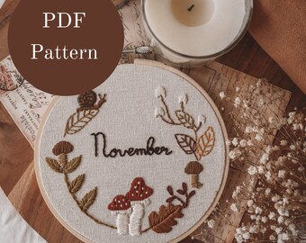 May include: Embroidered hoop art with the word "November" surrounded by mushrooms, leaves, and a snail. The hoop is on a wooden surface with white flowers.