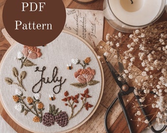 May include: Embroidered hoop art with a floral design and the word "July" in cursive. The hoop is on a wooden surface with a candle, a glass jar with dried flowers, and a pair of scissors.