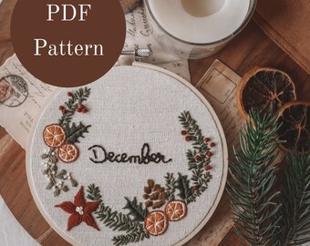 May include: A white embroidery hoop with a hand-stitched design of a wreath with oranges, pine needles, berries, and a poinsettia. The wreath surrounds the word "December" in black cursive lettering.